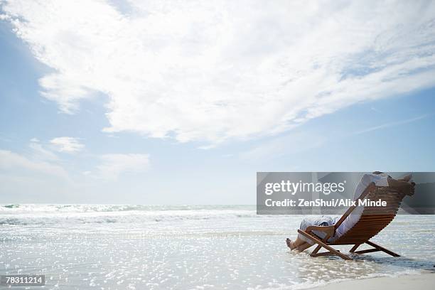 man sitting in deckchair on beach, surf washing under him, rear view - man on the beach relaxing in deckchair fotografías e imágenes de stock