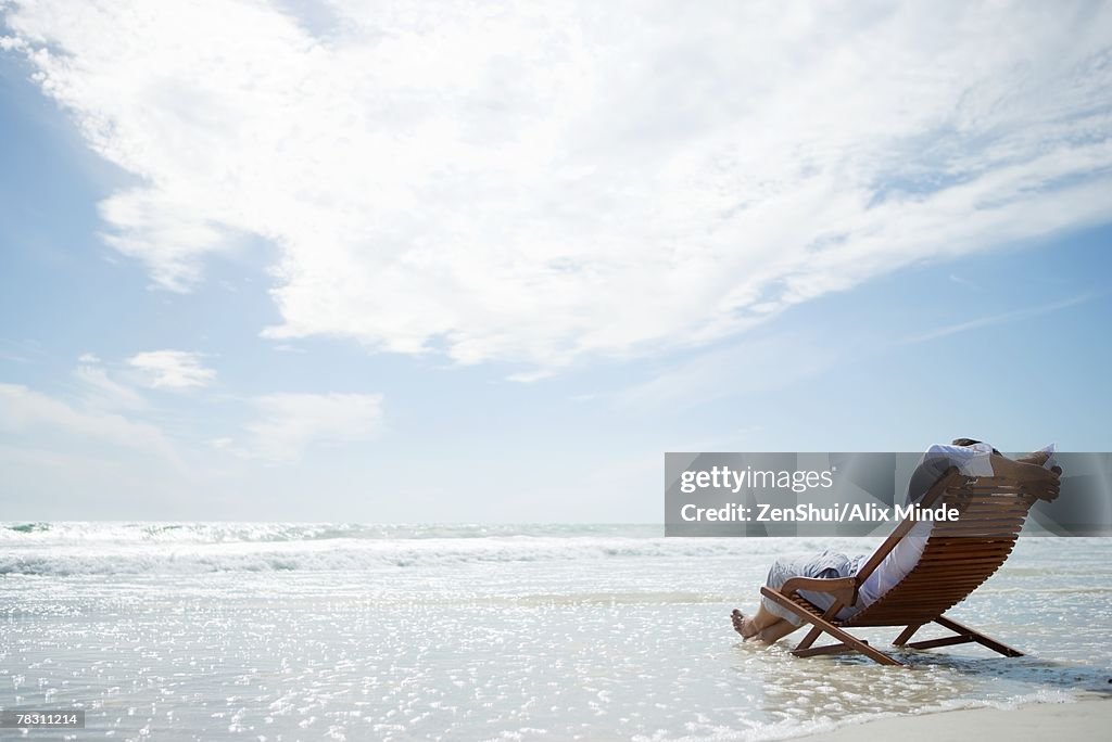 Man sitting in deckchair on beach, surf washing under him, rear view