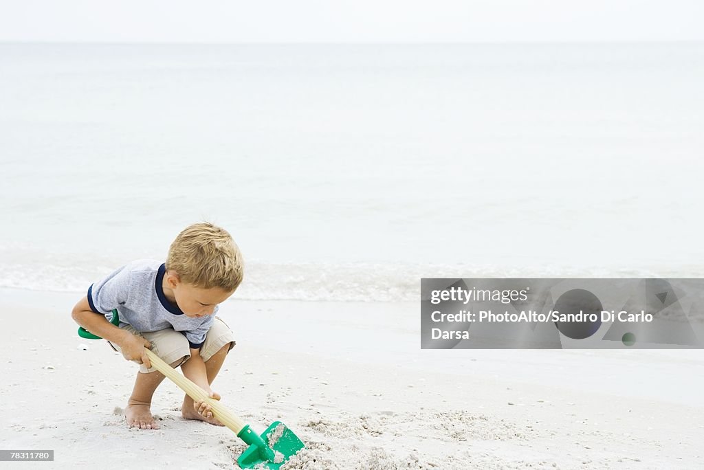 Young boy crouching at the beach, digging in sand with shovel