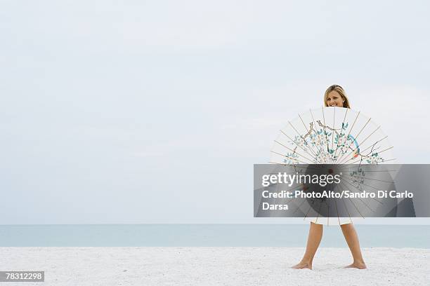 woman walking behind parasol at the beach, smiling at camera - papierschirm stock-fotos und bilder