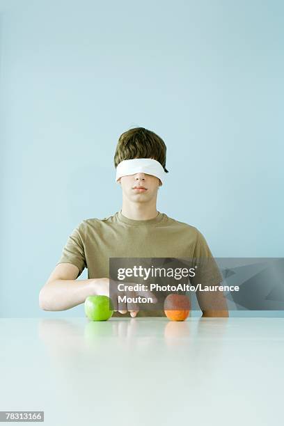 young male wearing blindfold, pointing to one of two apples - blinddoek stockfoto's en -beelden