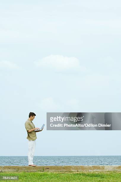 man standing on low wall using laptop, body of water in background, full length - man standing full body photos et images de collection