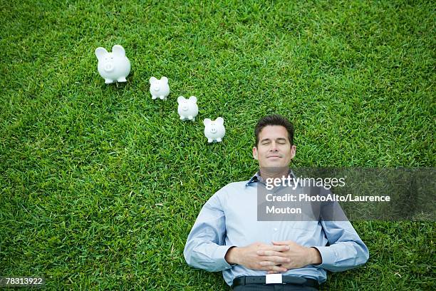 man lying on grass, line of piggy banks by head - planalto stock pictures, royalty-free photos & images