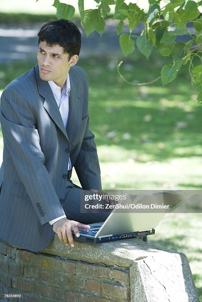 Businessman sitting on low wall in park, using laptop, looking away