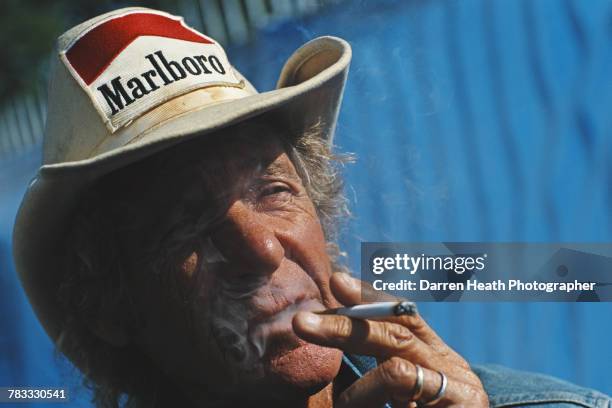 Former F1 Grand Prix and Sportscar driver Arturo Merzario, smokes a cigarette whilst wearing his trademark cowboy hat with sponsorship patches from...