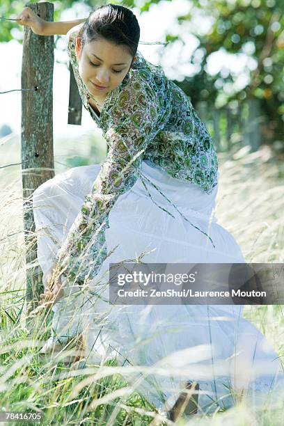 young woman holding onto fence, bending over to pick sprig of grass - bending over in skirt stock-fotos und bilder