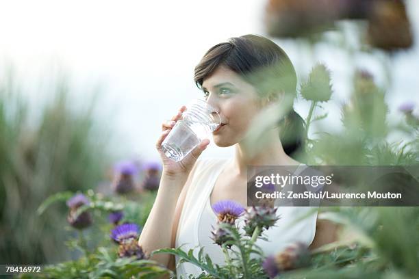 young woman drinking glass of water outdoors, surrounded by thistle flowers - water and flowers stock-fotos und bilder