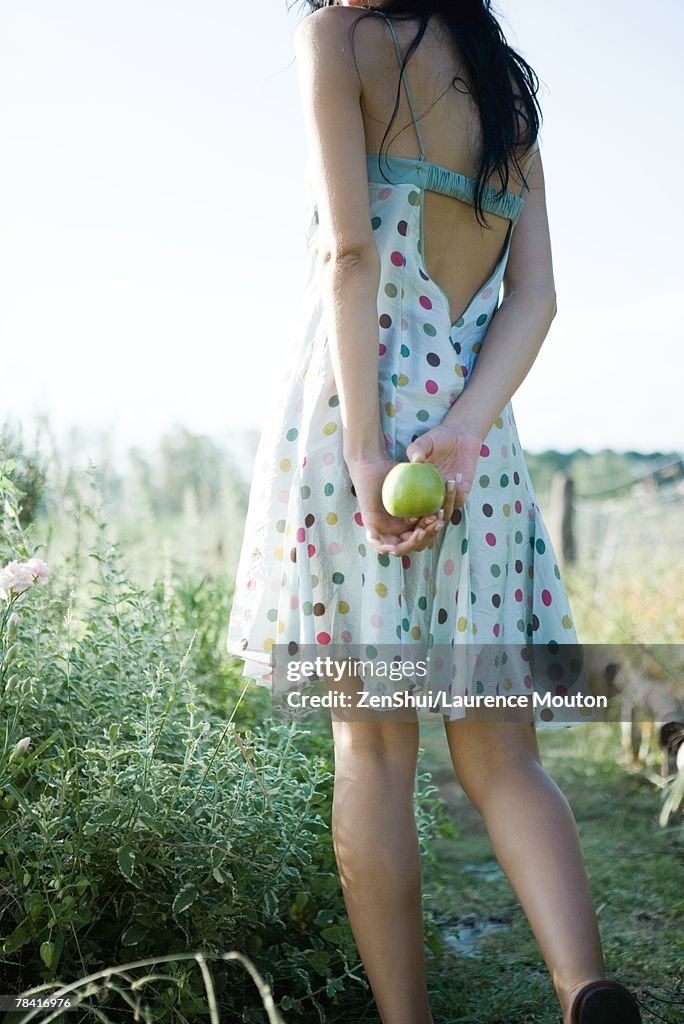 Young woman standing outdoors, holding apple behind back, cropped