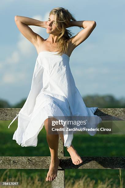 young woman sitting on top of wooden fence, hands behind neck, smiling - hands behind head stock pictures, royalty-free photos & images