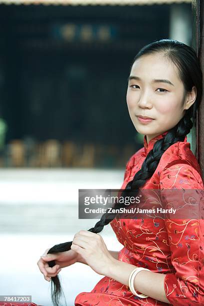 young woman dressed in traditional chinese clothing, holding her long braided hair, looking at camera - the cheongsam stock-fotos und bilder