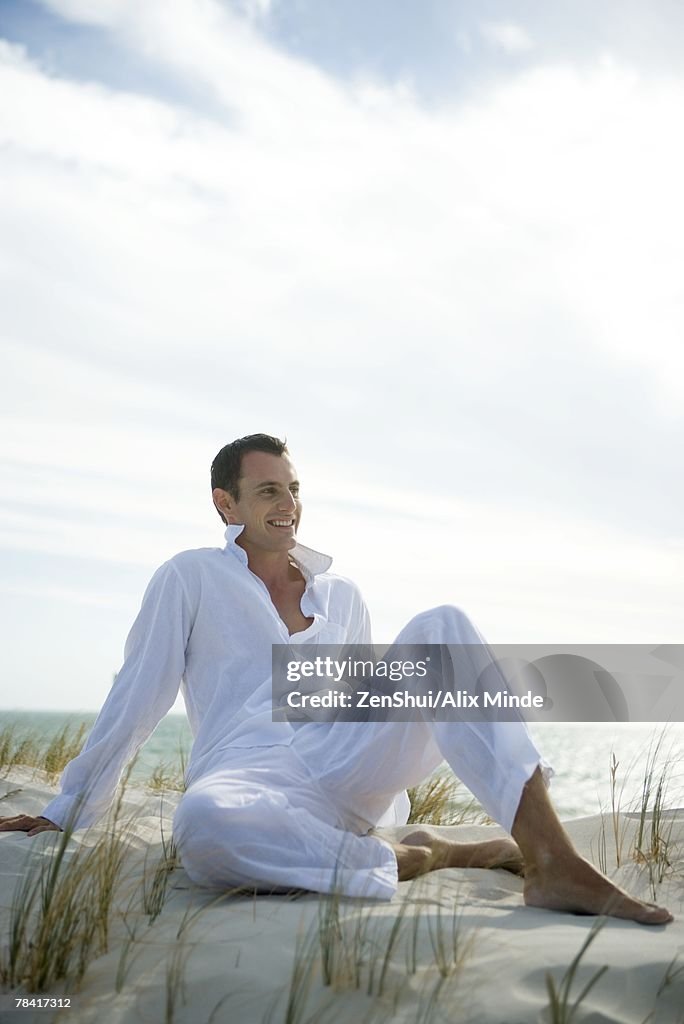 Young man sitting on dune, smiling, full length