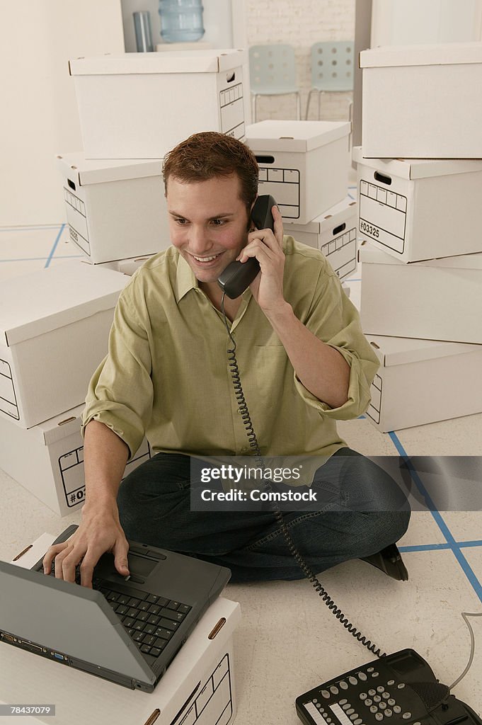 Man on telephone and using laptop with moving boxes behind him