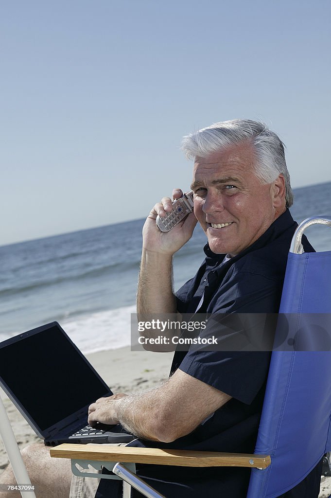 Man on cell phone and using laptop computer at beach