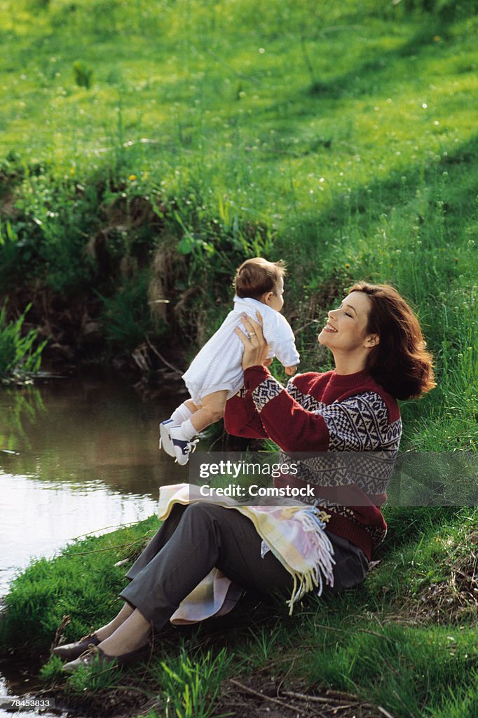 Mother sitting at lake's edge holding up baby