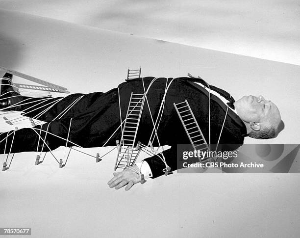 Promotional portrait of British-born American film and television director Alfred Hitchcock as he lies, tied to pegs in the floor and covered with...