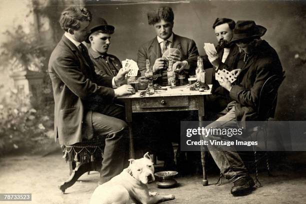 Staged portrait of five men as they play poker crowded around a small table cluttered with beer bottles, glasses, and handguns, Rush City, Minnesota,...