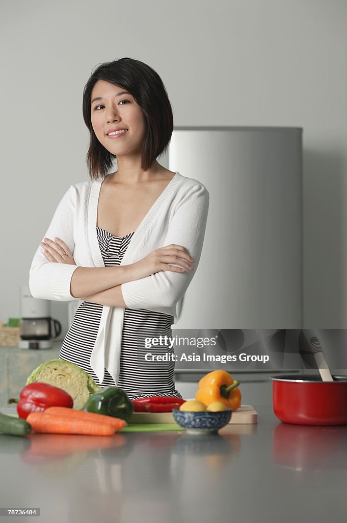 Young woman in kitchen, arms crossed, vegetables on the table in front of her