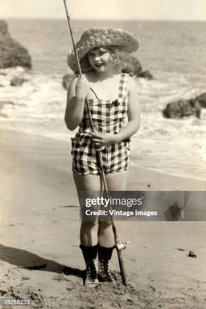 Portrait of an unidentified young woman, one of Mack Sennett's 'bathing beauties,' who poses at the beach in a straw hat, checkered bathing costume,...