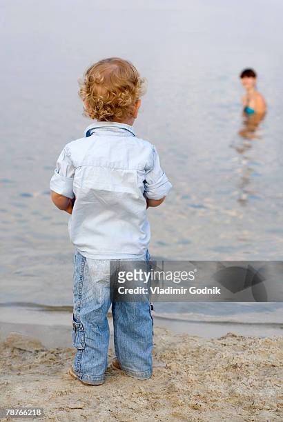 a young boy standing on a beach and looking at a woman swimming - boy curly blonde stock pictures, royalty-free photos & images