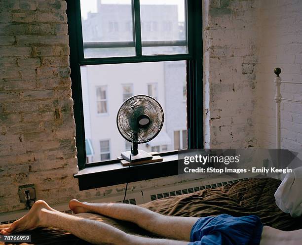 a man lying on a bed with a fan blowing - caldo foto e immagini stock