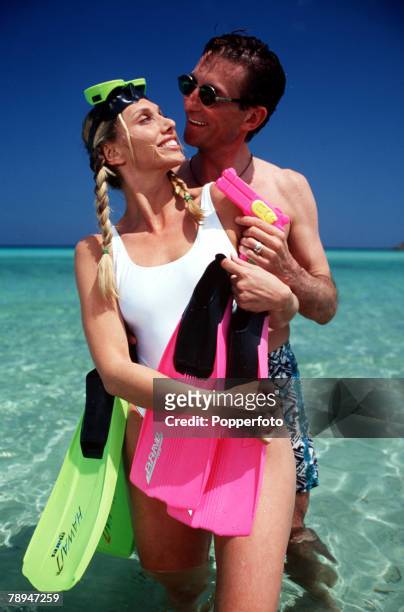 Stock Photography, Young couple standing together in clear tropical water, carrying brightly coloured flippers and posing playfully with a pink water...