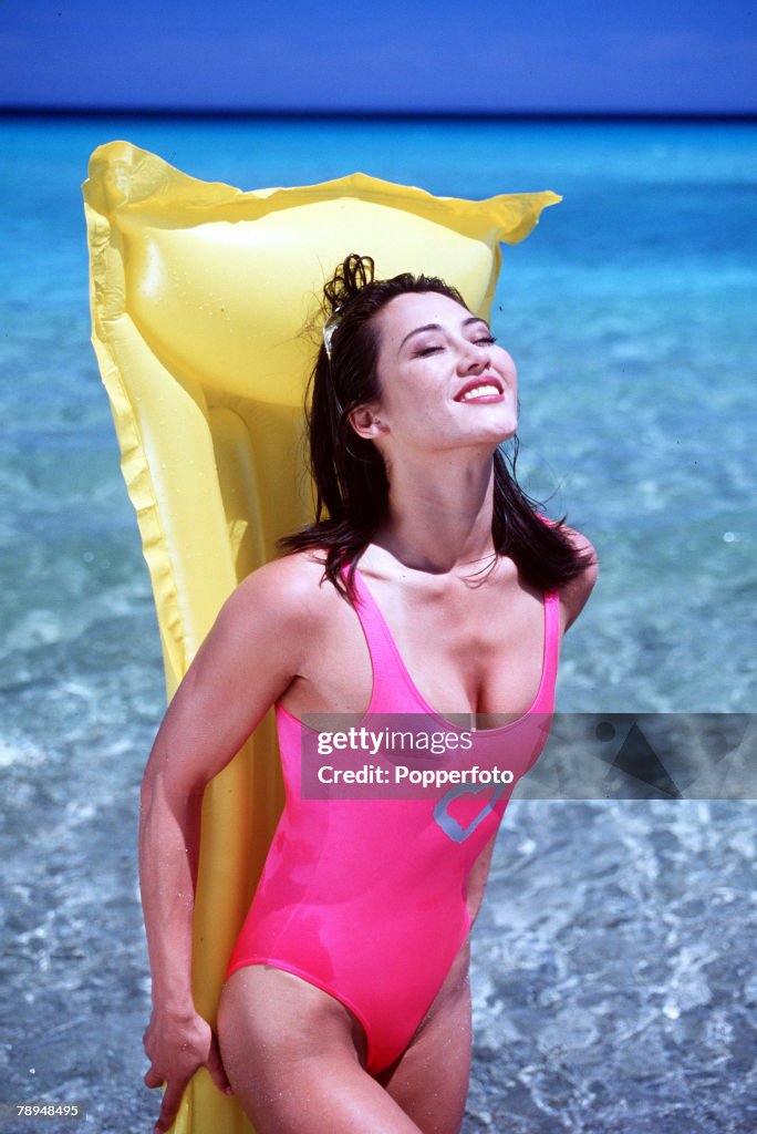 Stock Photography. Young glamourous woman wearing bright pink swimming costume, stands at the water+s edge with a yellow lilo held tight against her back as she enjoys the warm sunshine.