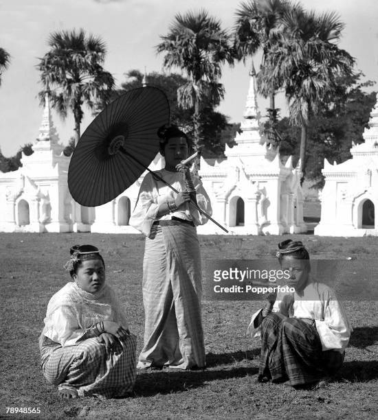 Ponting in Asia 1900 - 1906, Burma, A Native girl in traditional dress stands holding a parasol and smoking a cheroot with two other girls crouching...
