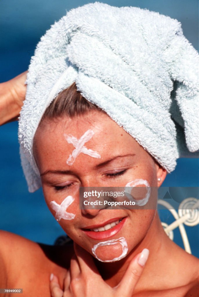 Stock Photography. Portrait of a young woman relaxing in the sunshine with skin cream smeared in a pattern of noughts & crosses on her face and wearing a towel wrapped around her hair.