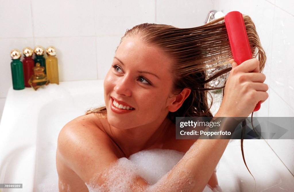 Stock Photography. Young smiling woman sitting up in a bath as she pulls a brush through her wet hair.