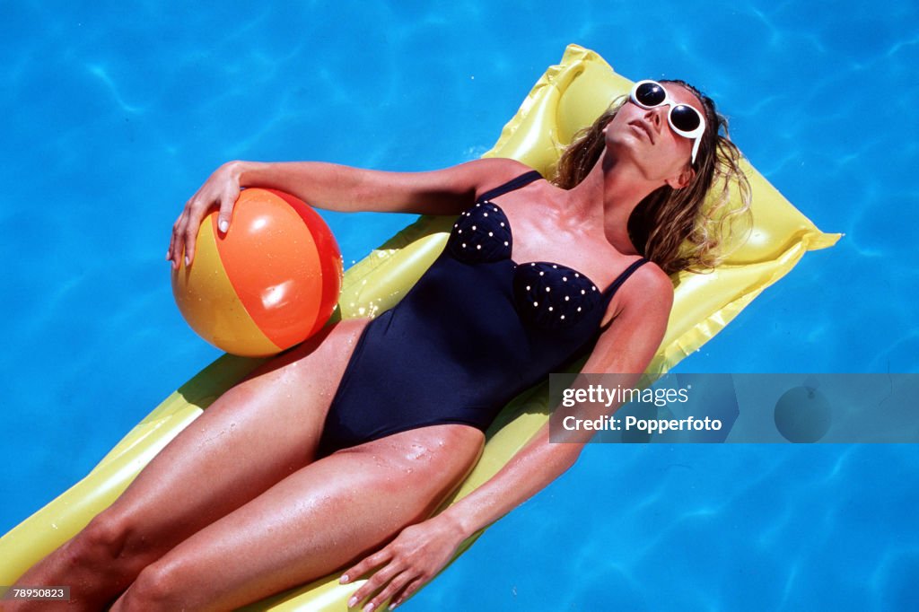 Stock Photography: Tanned woman wearing swimming costume and sunglasses, floating on a lilo in a pool, and clutching a beach ball. Overhead view looking down.