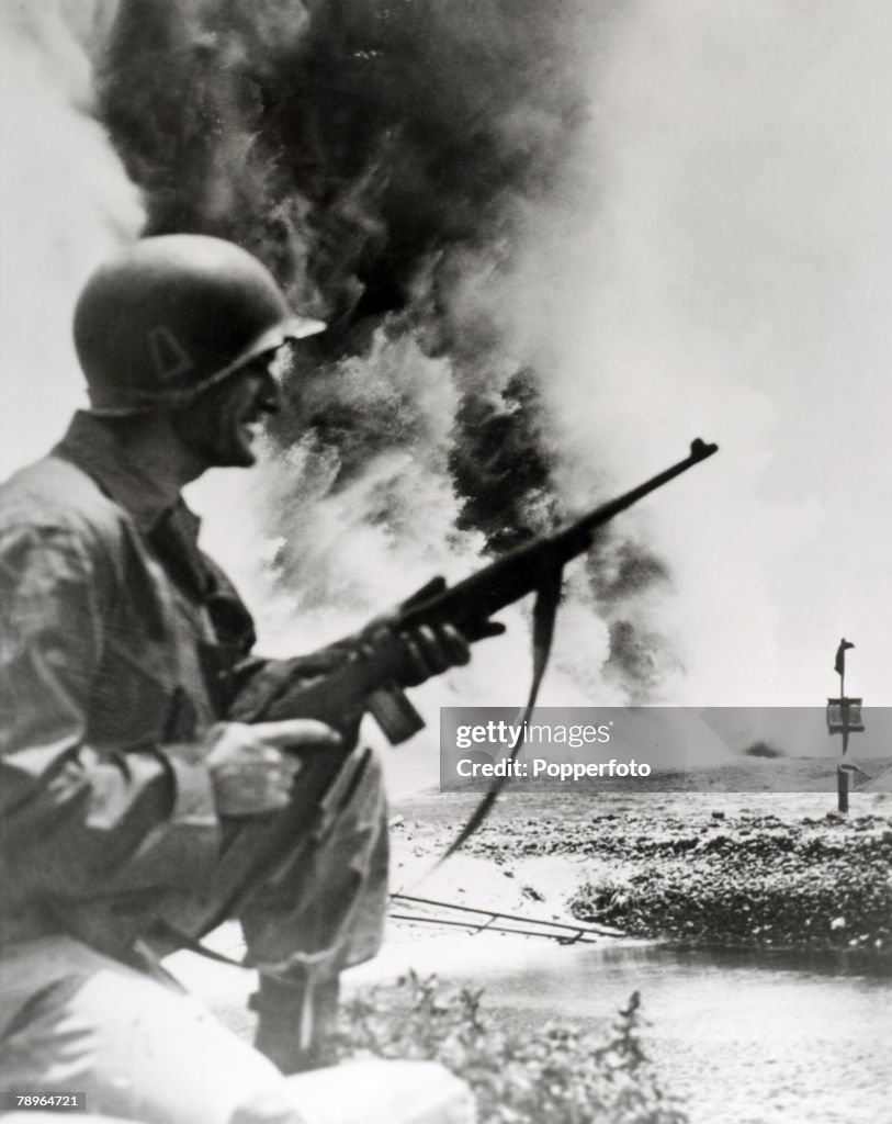 War and Conflict. World War Two. 1945. Okinawa, Pacific. An American soldier holds his rifle with explosions and smoke in the background.