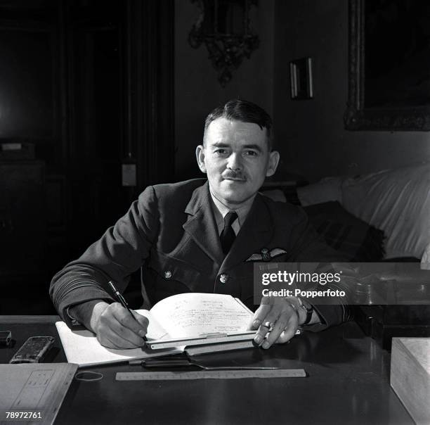 World War II, Group Captain Frank Whittle wearing military uniform of the Royal Air Force as he sits at his desk writing in a note book with the aid...