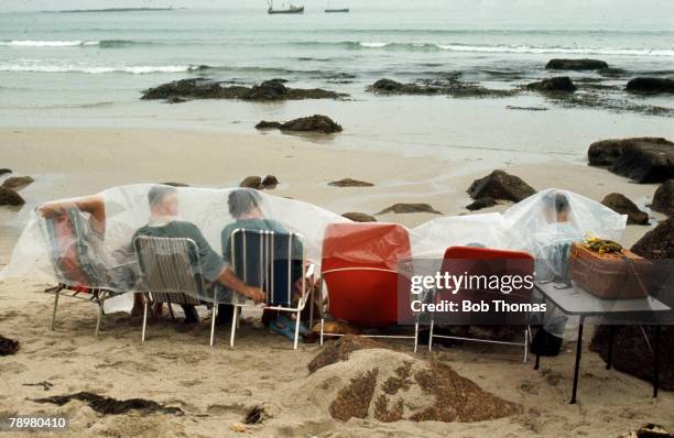 People, Holidays, England, pic: 1970, Hardy holidaymakers hoping for a picnic have to endure the British climate as they take shelter on the beach by...