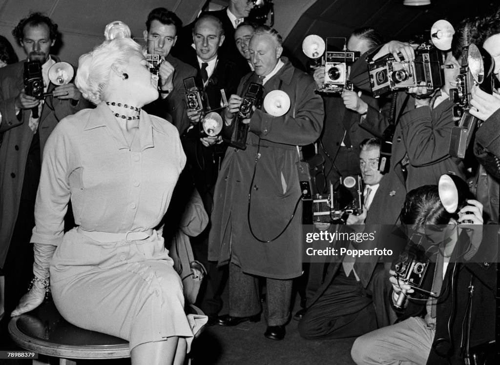 PF Stage & Screen. London, England. 1957. American film and movie actress Jayne Mansfield sits on a small table surrounded by photographers and reporters, while attending a press conference.