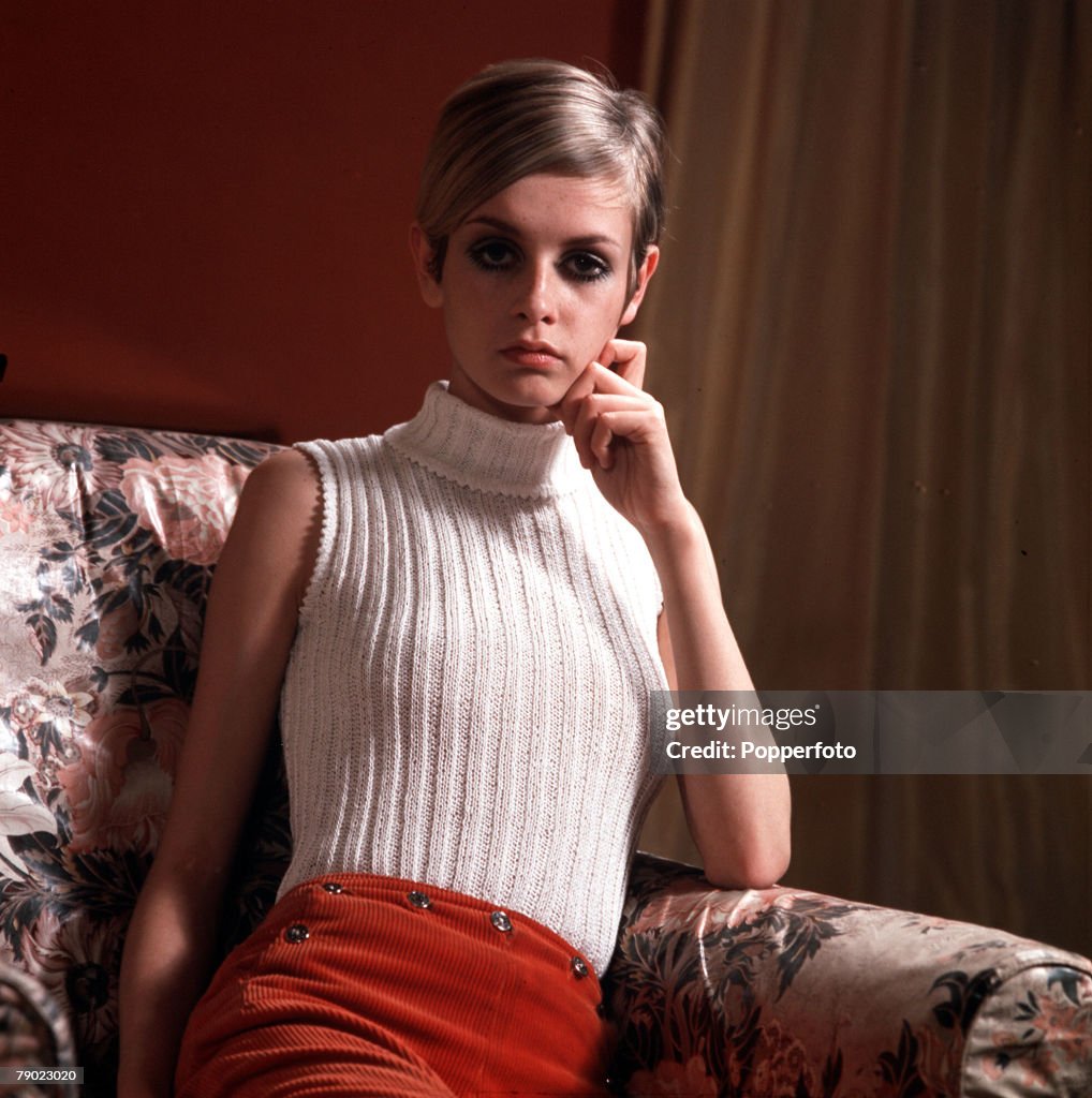 1967. Modelling. A picture of British model Twiggy wearing a fashionable sleeveless, beige polo neck jumper and red trousers, as she poses on a settee.