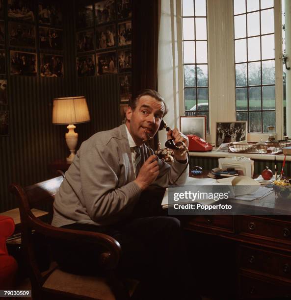 Picture of the British actor Ian Carmichael sitting at a desk near a window whilst on the telephone looking towards the camera