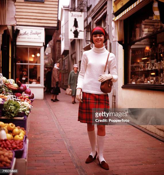 Young woman wearing a white polo neck top and red tartan skirt standing in front of a shop selling fruit and vegetables