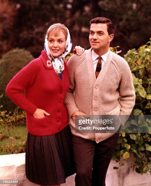 Female and male fashion model wearing coloured woollen cardigans in red and beige, they stand together outside in a garden area, 1961.