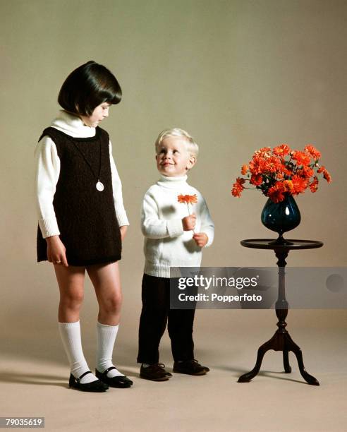 Children+s Fashion 1964: Smiling young boy wearing a white polo neck sweater holds an orange flower that he has removed from a vase, as an older girl...