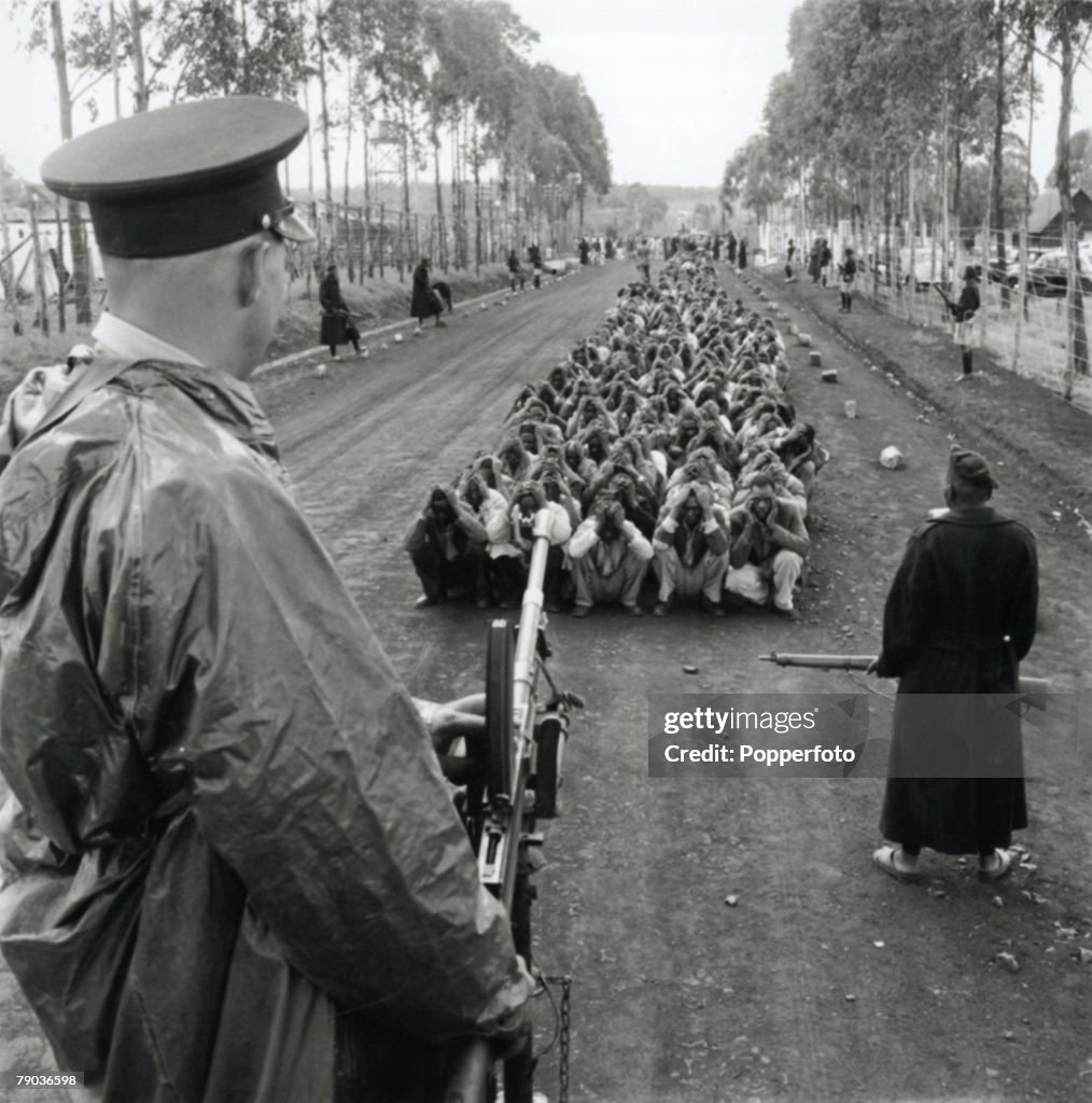 Nairobi, Kenya, 24th April, 1954. A policeman stands guard with a gun over tribesmen, suspected of committing Mau Mau crimes.