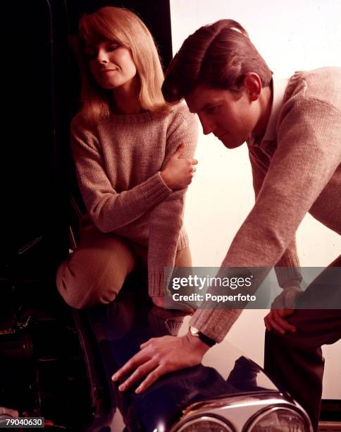 Posed studio image of a man peering under the bonnet of a car while a woman sits on the wing and looks in too, they both wear matching oatmeal...