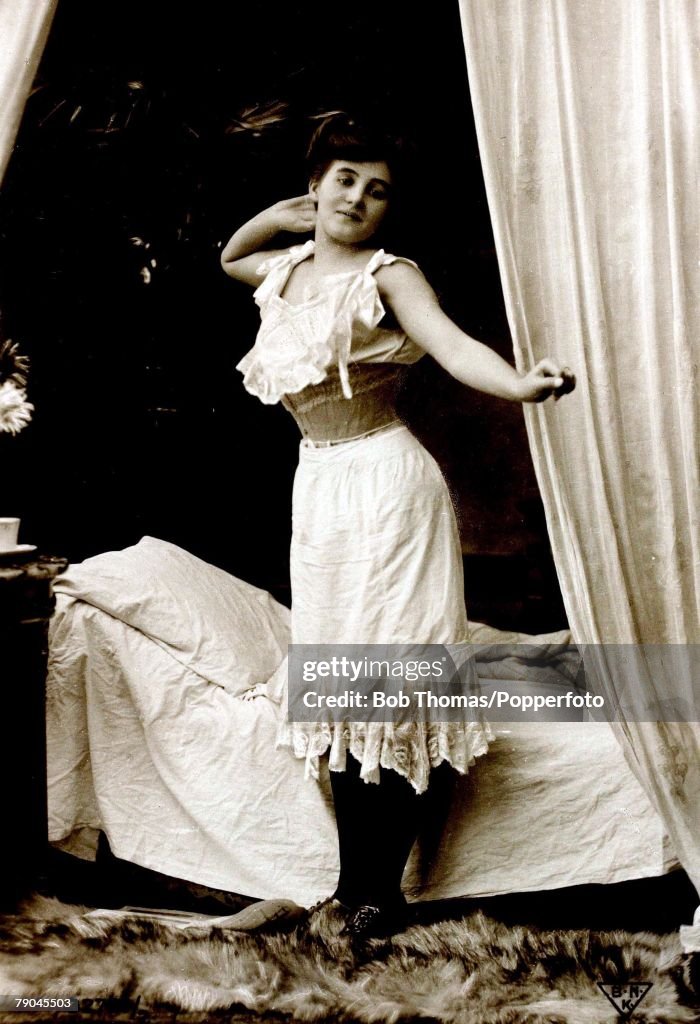 Postcards. Circa 1915. Dishabille. A picture of a woman wearing an undergarment and corset whilst standing by a bed posing for the camera.