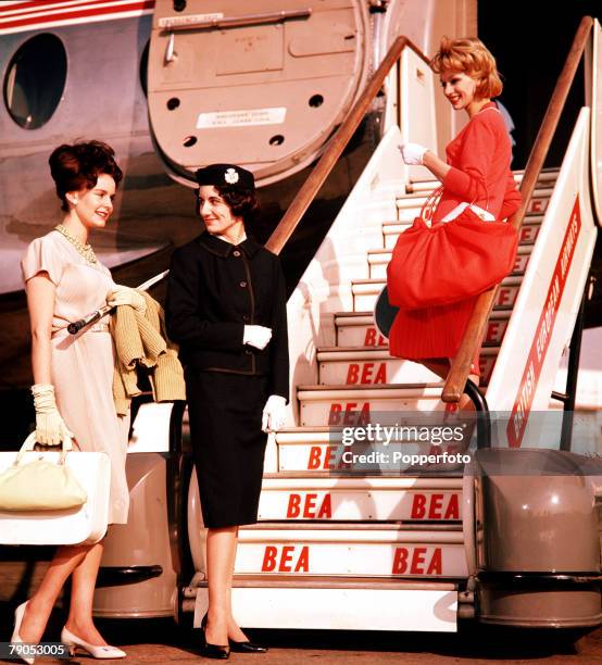 Two female fashion models walking up a set of steps to an airplane, with a smiling stewardess, at an airport in England, 1960.