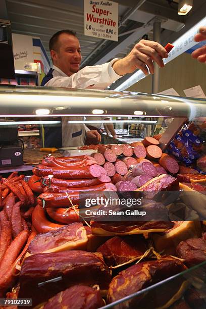 Butcher offers a visitor a sample of dried sausage as different types of traditional German sausage, ham, salami and other meat products lie on...