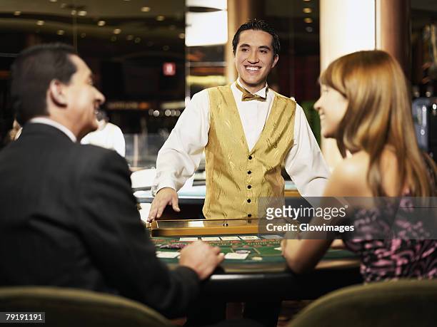 rear view of a mature man and a young woman sitting at a gambling table with a casino worker smiling in front of them - croupier foto e immagini stock