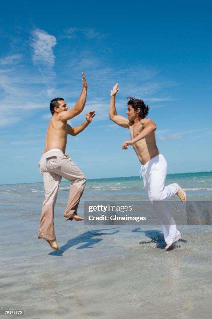 Two mid adult men jumping and giving high-five to each other on the beach