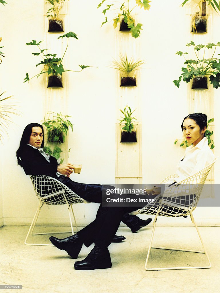 Young couple sitting on wire-framed chairs, potted plants decorated wall background