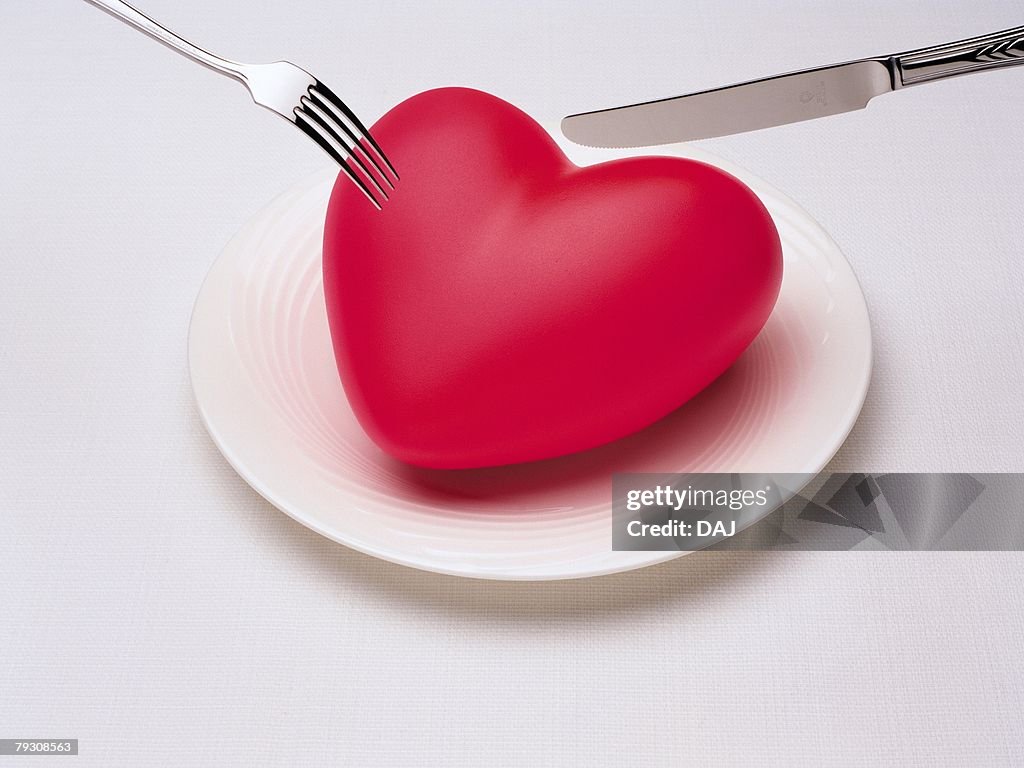 Fork and knife reaching to red heart on plate, high angle view, white background