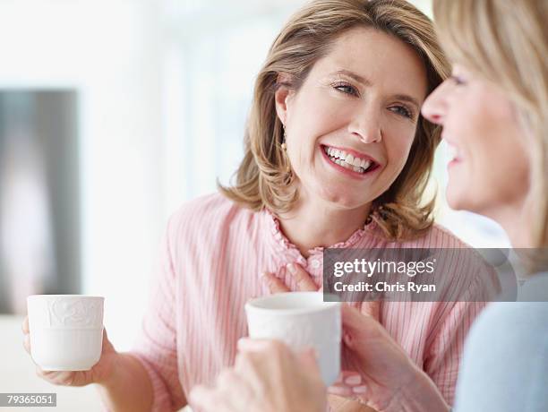 two women indoors holding mugs and talking - mature women coffee stock pictures, royalty-free photos & images