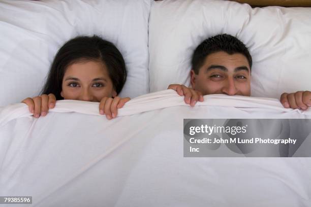 multi-ethnic couple holding bed sheet over mouths - romantic young couple sleeping in bed stock pictures, royalty-free photos & images